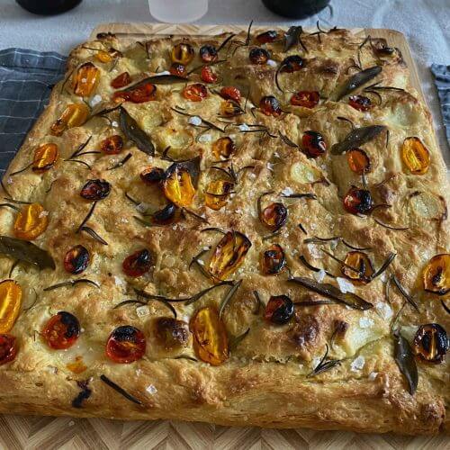 A freshly baked Italian focaccia topped with fresh sage leaves, sprigs of rosemary, and ripe tomatoes.