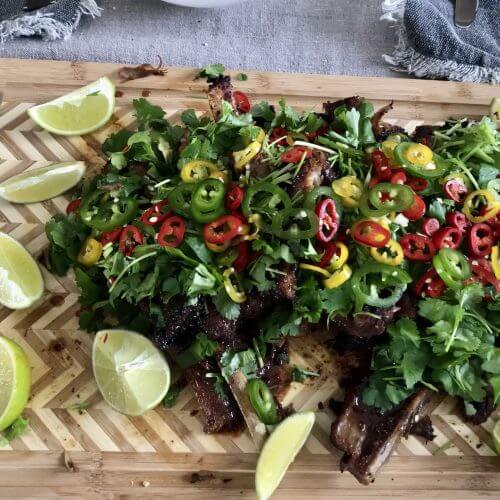Pork spareribs on a wooden cutting board, topped with fresh cilantro and colorful chopped chilies