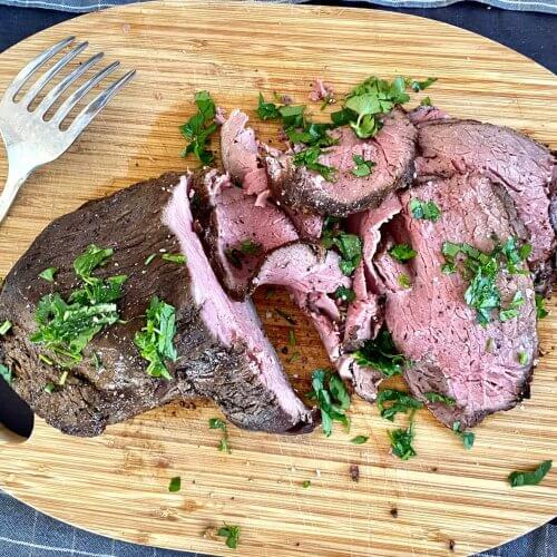 Juicy marinated roast beef sliced on a wooden cutting board, garnished with fresh chopped parsley.