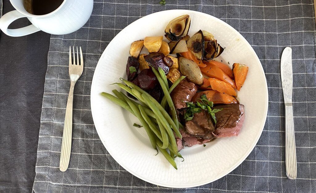 Sliced roast beef topped with gravy on a plate, served with green beans, roasted carrots, ruabaga, onions, and beets.