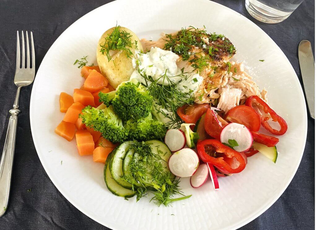 Grilled salmon served with new potatoes, horseradish-kermaviili sauce, fresh mixed salad, steamed carrots, and broccoli on a white plate.