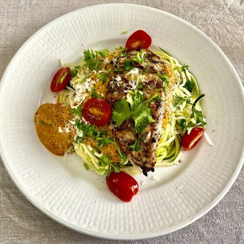 Grilled chicken with cashew romesco sauce and zucchini noodles, garnished with sweet cherry tomatoes and fresh parsley.