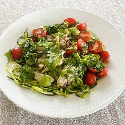 A plated summer dish featuring grilled chicken breast, zucchini spaghetti, cherry tomatoes, cashew pesto, and fresh basil leaves.
