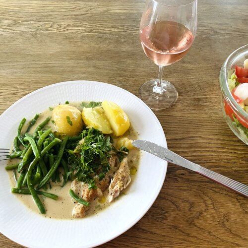 White plate with sage chicken and new potatoes, served with a glass of rosé wine and a side of mixed salad.
