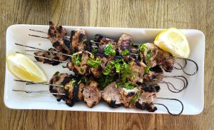 Grilled pork souvlaki skewers on a white plate, garnished with fresh lemon wedges.