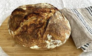 Freshly baked Dutch oven bread on a wooden cutting board with a linen kitchen towel beside it.
