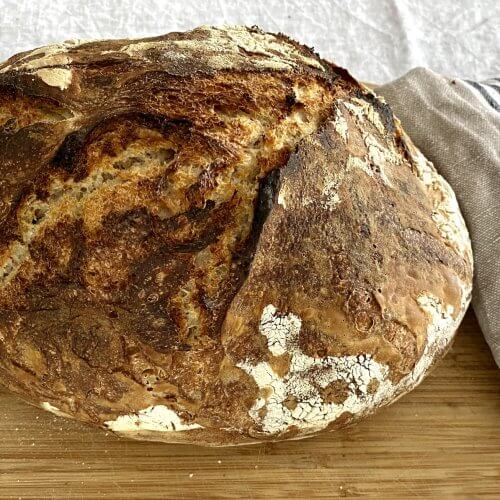 Freshly baked Dutch oven bread on a wooden cutting board with a linen kitchen towel beside it.