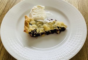 A slice of rustic blueberry pie served on a white plate with a scoop of melting vanilla ice cream beside it.