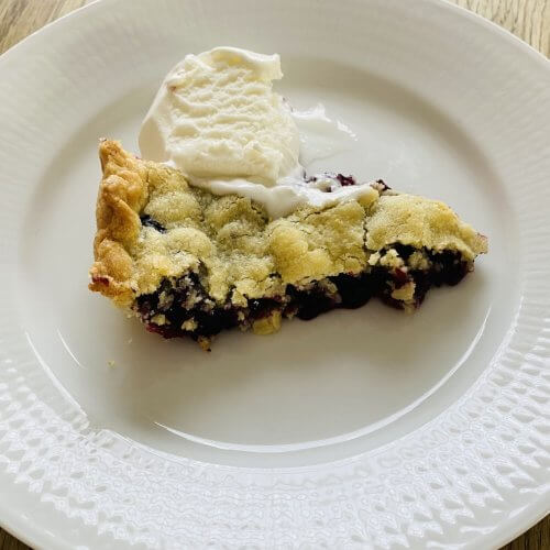A slice of rustic blueberry pie served on a white plate with a scoop of melting vanilla ice cream beside it.