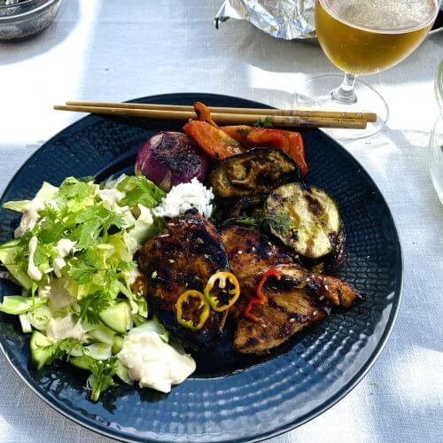 Grilled teriyaki chicken with vegetables and mixed green salad on a dark grey plate, garnished with chili and cilantro. Chopsticks rest neatly on the plate's edge.