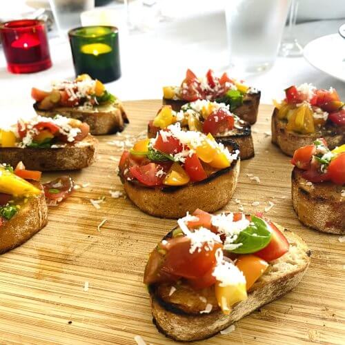 Tomato-basil bruschettas with grated Parmesan Reggiano on a wooden cutting board, set on a white linen tablecloth with colorful candle holders in the background.