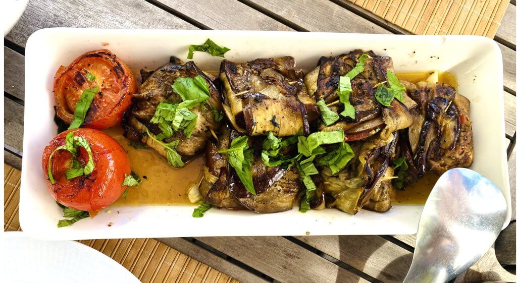 Grilled eggplant bundles stuffed with mozzarella and basil, served with grilled whole tomatoes on a white plate