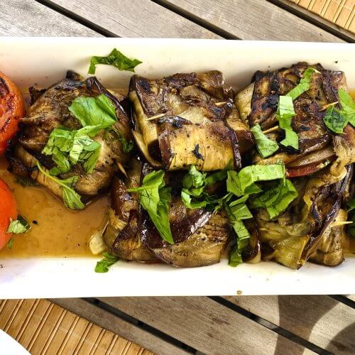 Grilled eggplant bundles stuffed with mozzarella and basil, served with grilled whole tomatoes on a white plate