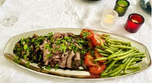 Sliced grilled beef sirloin roast on a serving platter with grilled vegetables, fresh herbs, and colorful candle holders on a white linen tablecloth