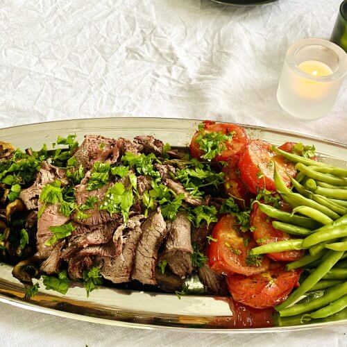Sliced grilled beef sirloin roast on a serving platter with grilled vegetables, fresh herbs, and colorful candle holders on a white linen tablecloth