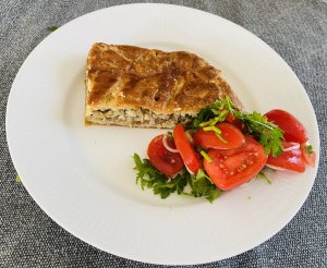 A slice of Finnish slab meat pie (peltilihapiirakka) on a white plate, served with a fresh tomato and herb salad.