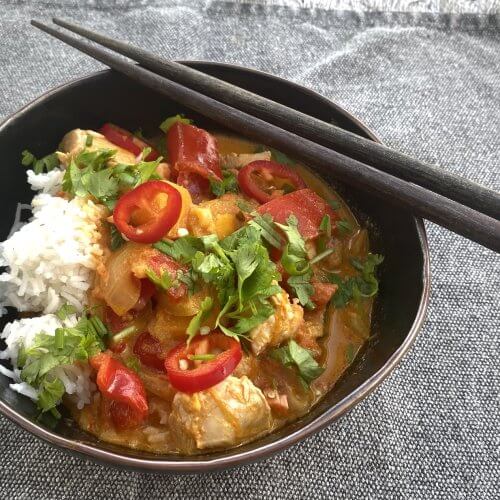 A rustic bowl filled with Thai-style red curry, garnished with fresh red chili slices and chopped cilantro, served hot with steamed rice.