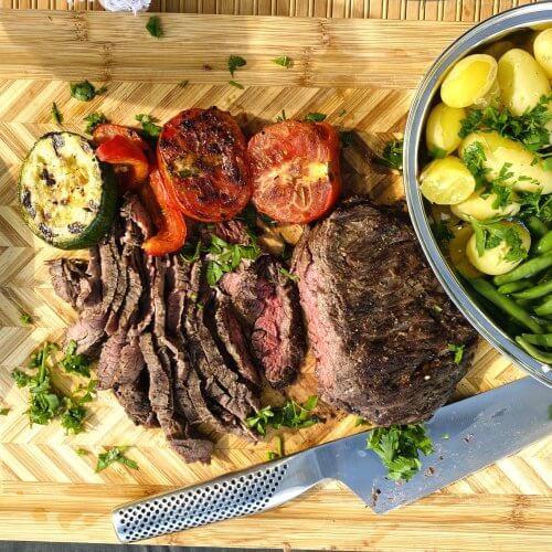 Thinly sliced medium-grilled beef flank steak on a wooden cutting board with grilled zucchini, red bell peppers, and tomato halves, alongside steamed new potatoes and green beans on a steel plate.