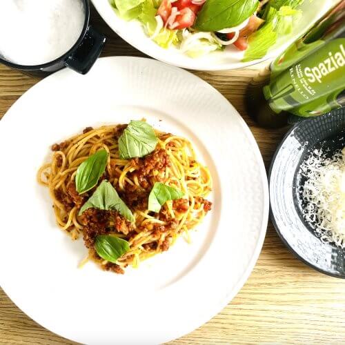 Plate of spaghetti Bolognese topped with grated parmesan and fresh basil, served alongside a bottle of olive oil, a small plate of grated parmesan, mixed salad, and a cup of flaky sea salt.