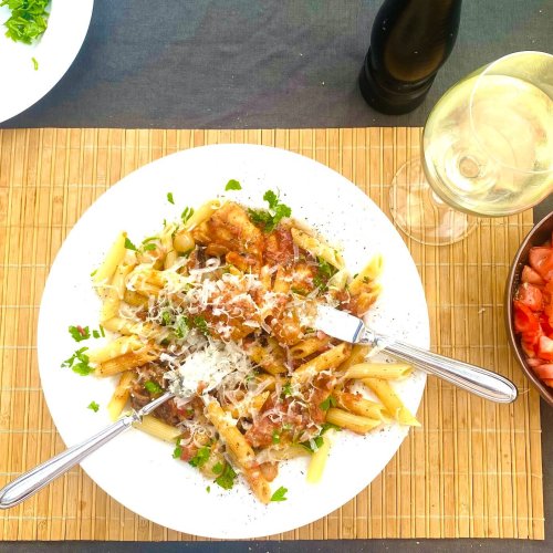 Chicken pasta with tomato, mushrooms, and white wine sauce, served on a white plate, accompanied by a glass of white wine, tomato salad, and grated Parmesan on the side, on a placemat.