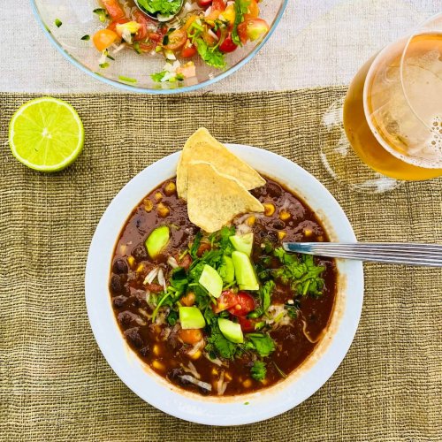 Vegetarian black bean and corn soup in a white bowl, topped with avocado, tomato-onion mix, cilantro, and tortilla chips, served with a side bowl of fresh tomato mix, a glass of Mexican beer, and half a lime.