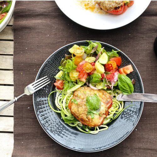 One half of a freshly baked red bell pepper stuffed with Bolognese, placed on a bed of zucchini spaghetti. A fresh side salad with colorful cherry tomatoes, cucumber, and basil is served alongside the plate.