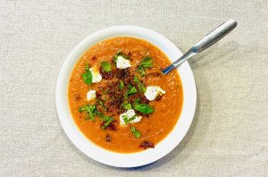 Roasted tomato soup served in a white bowl, topped with crispy bacon, fresh herbs, and cream cheese.