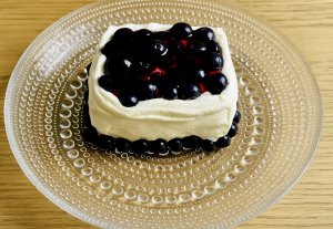 A blueberry mini cake on a glass plate, topped with a glossy blueberry glaze and surrounded by whipped cream, with fresh blueberries around the cake.