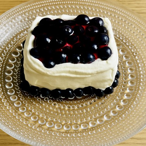 A blueberry mini cake on a glass plate, topped with a glossy blueberry glaze and surrounded by whipped cream, with fresh blueberries around the cake.