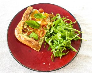 Slice of herbed cherry tomato tart on a red plate, served with fresh arugula salad.