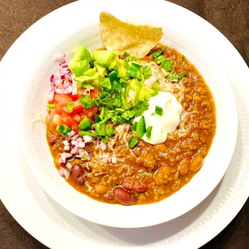 White bowl filled with hearty beef chili topped with sour cream, cilantro, diced red onions, tomato and avocado chunks, serrano slices, melted cheese and tortilla chips.