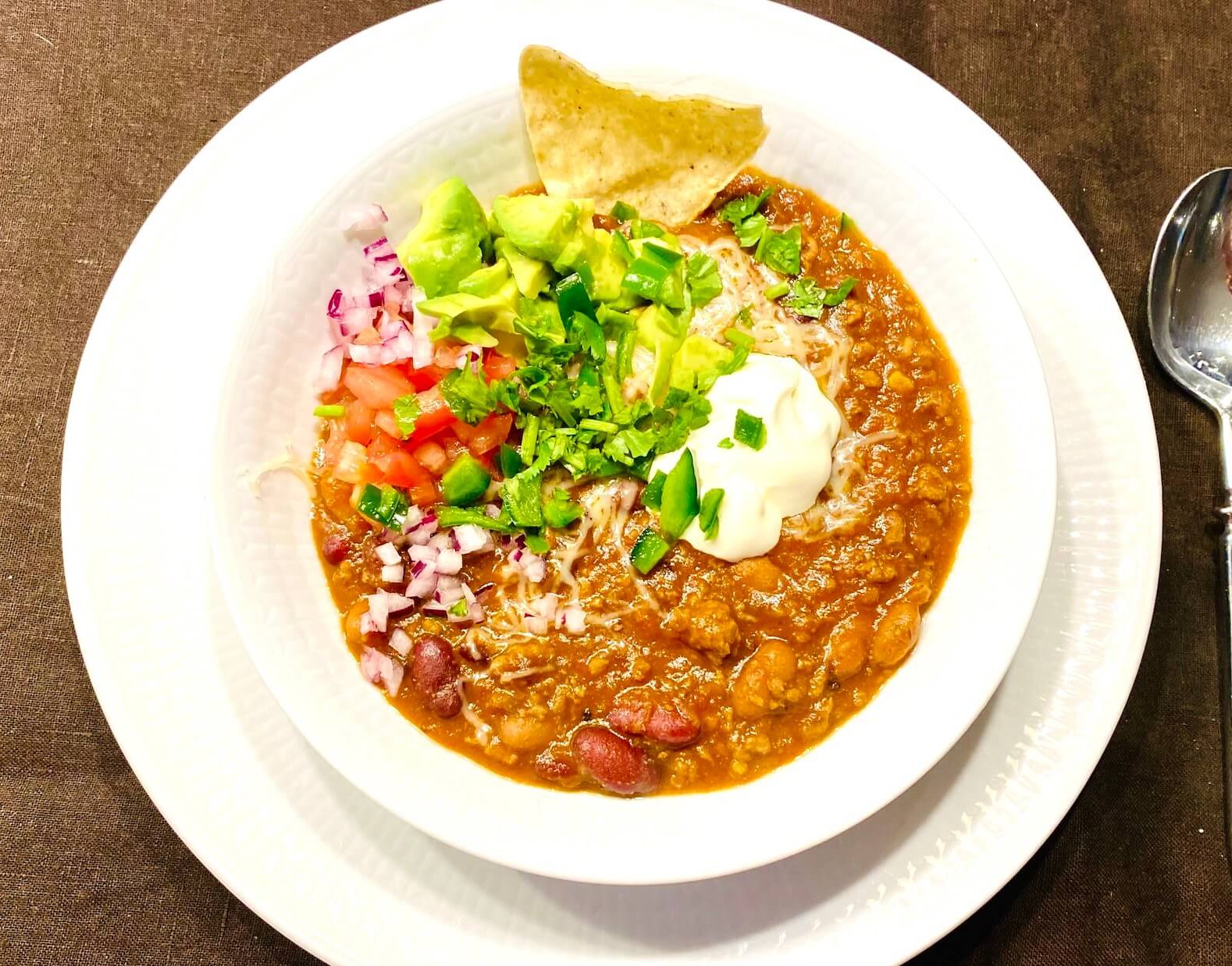 White bowl filled with hearty beef chili topped with sour cream, cilantro, diced red onions, tomato and avocado chunks, serrano slices, melted cheese and tortilla chips.