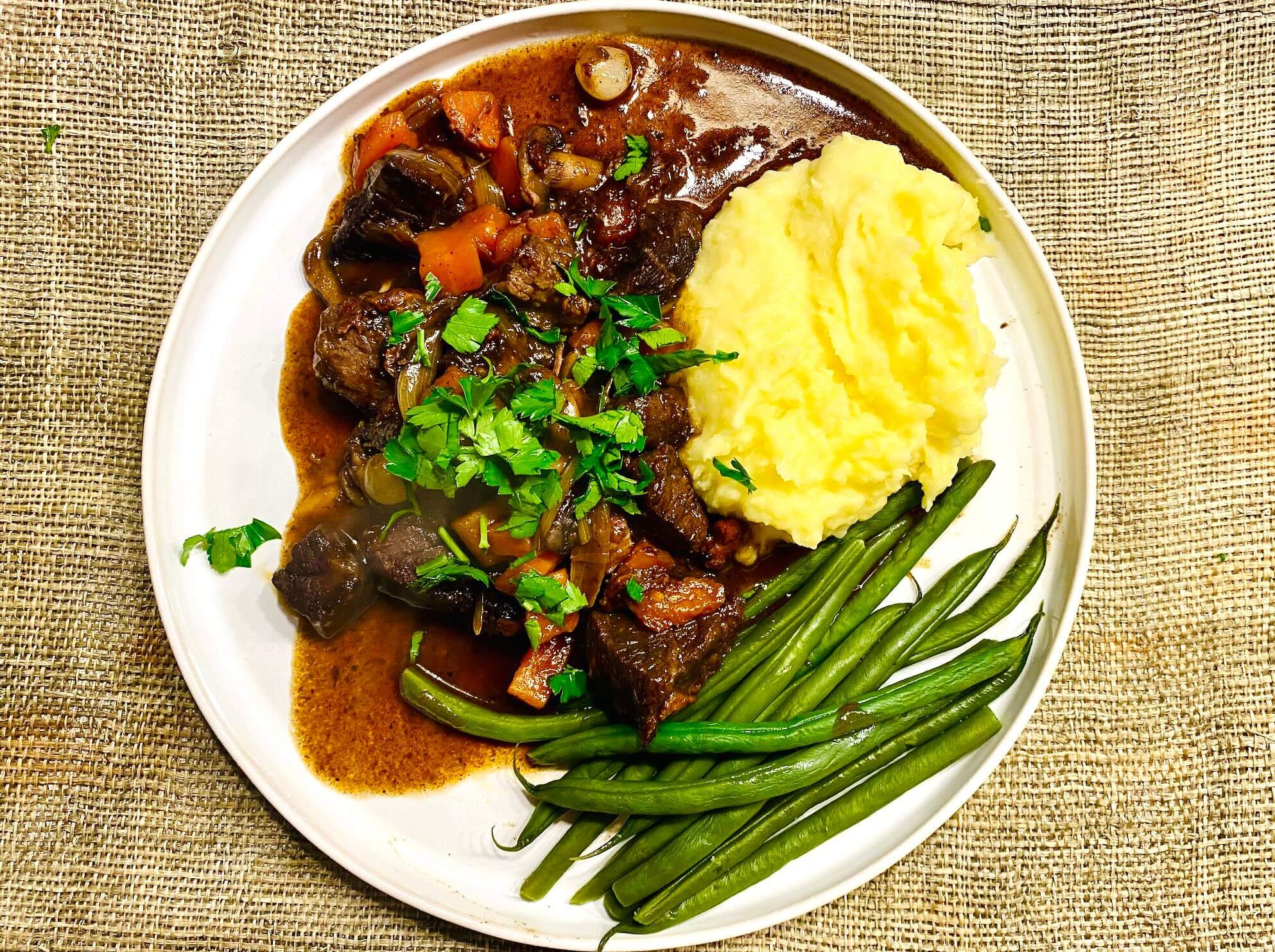 Beef Bourguignon with red wine sauce, served with creamy mashed potatoes and steamed green beans, garnished with fresh parsley.