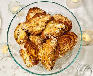 A pile of freshly baked Finnish cinnamon rolls, korvapuustit, arranged on a glass cake stand with candles around on a white tablecloth.