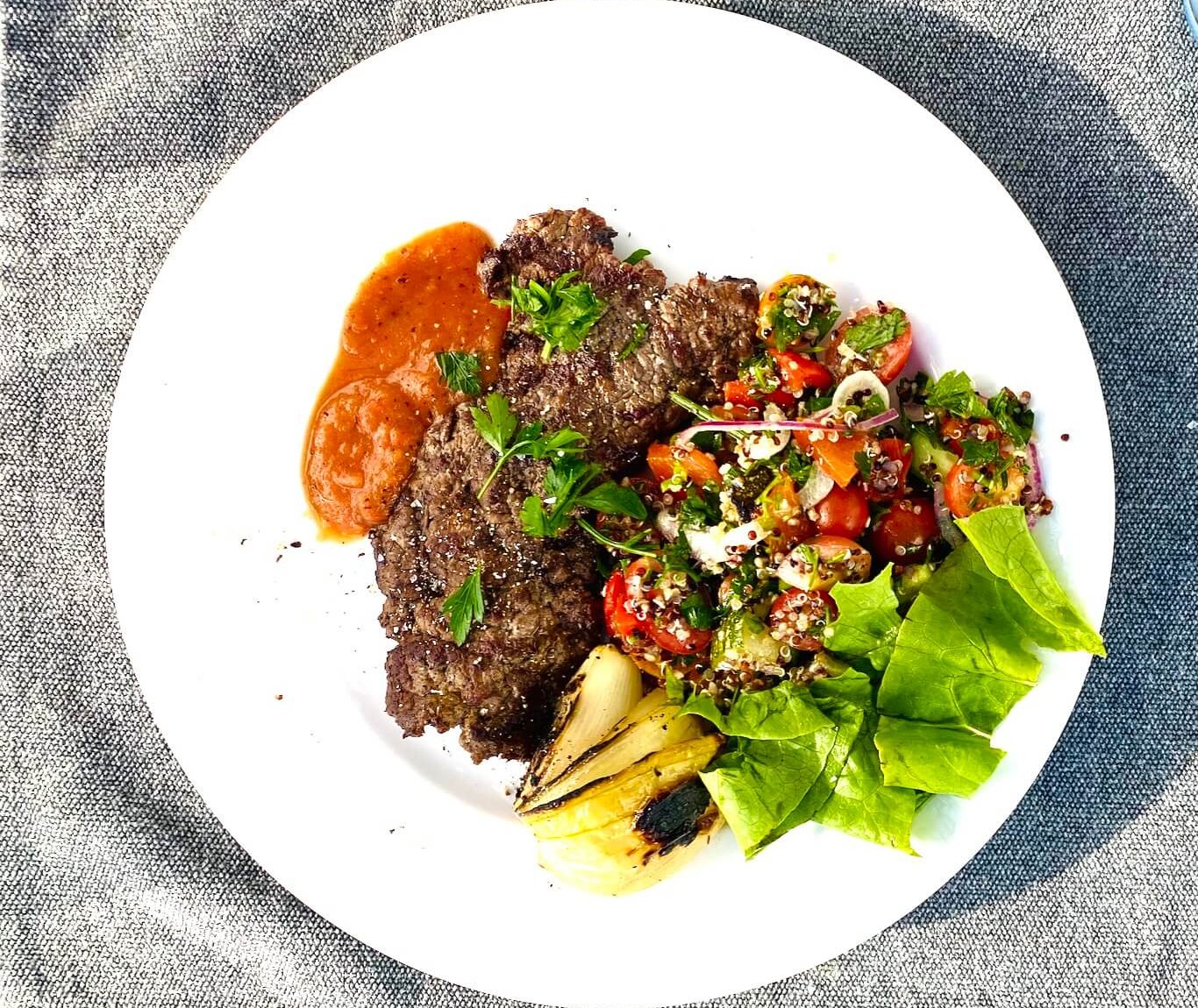 A plated Finnish-style thin beef steak with roasted red pepper sauce, grilled onion wedge, tomato-quinoa salad, and fresh lettuce.