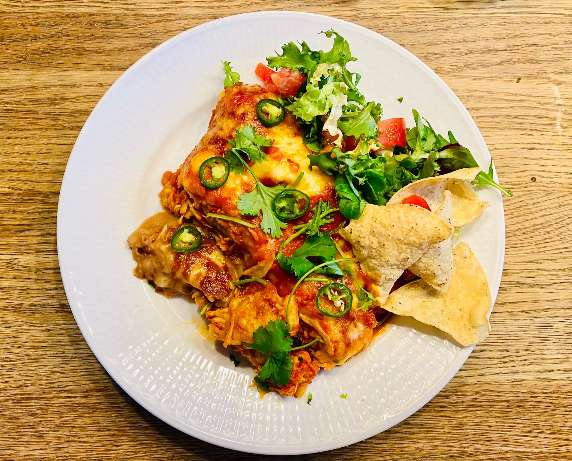Plate of chicken enchiladas with red chili sauce, served with salad, corn tortilla chips, fresh cilantro, and jalapeño slices.