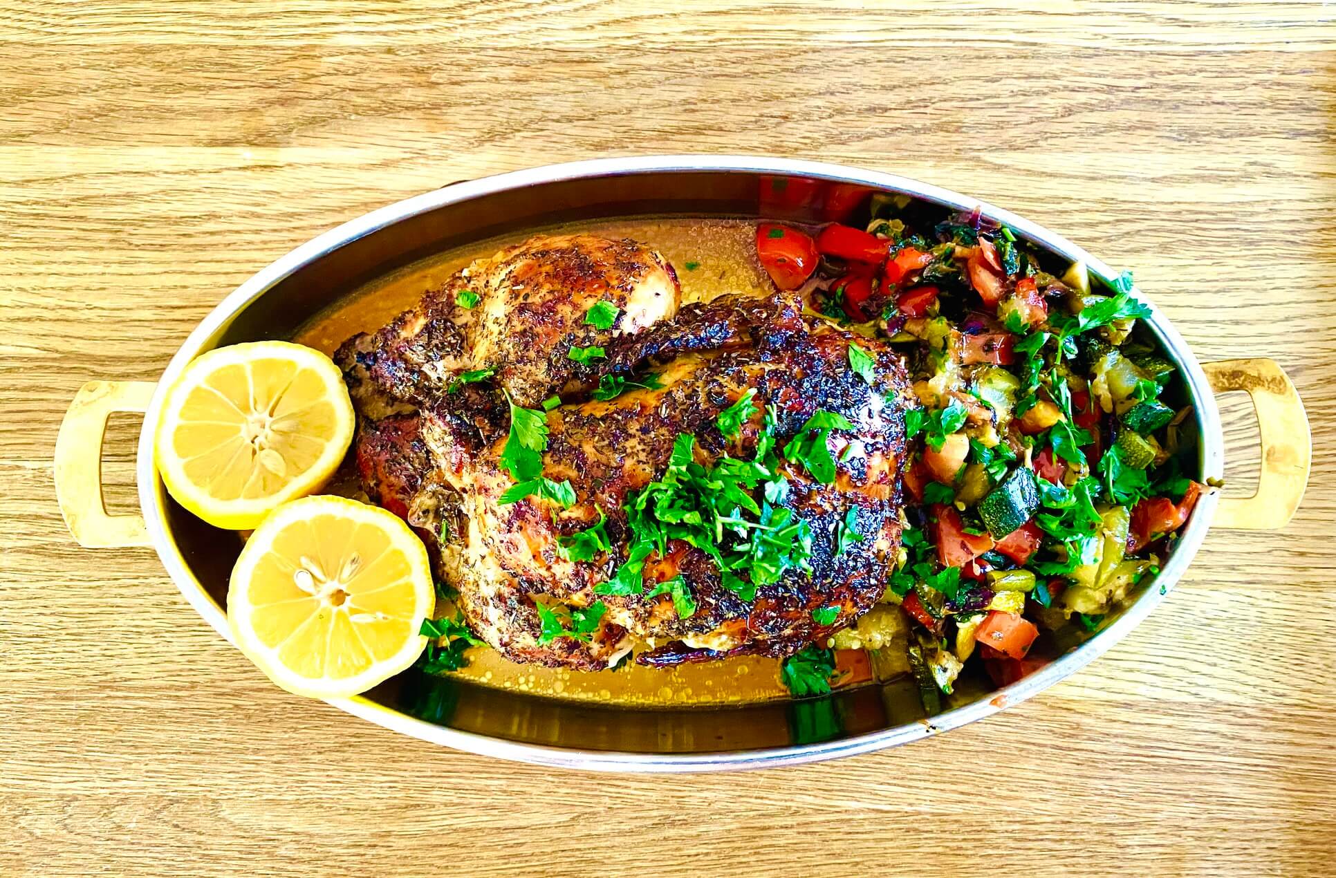 Oven-roasted whole chicken with vegetables, garnished with parsley and lemon halves, served in an oval dish.
