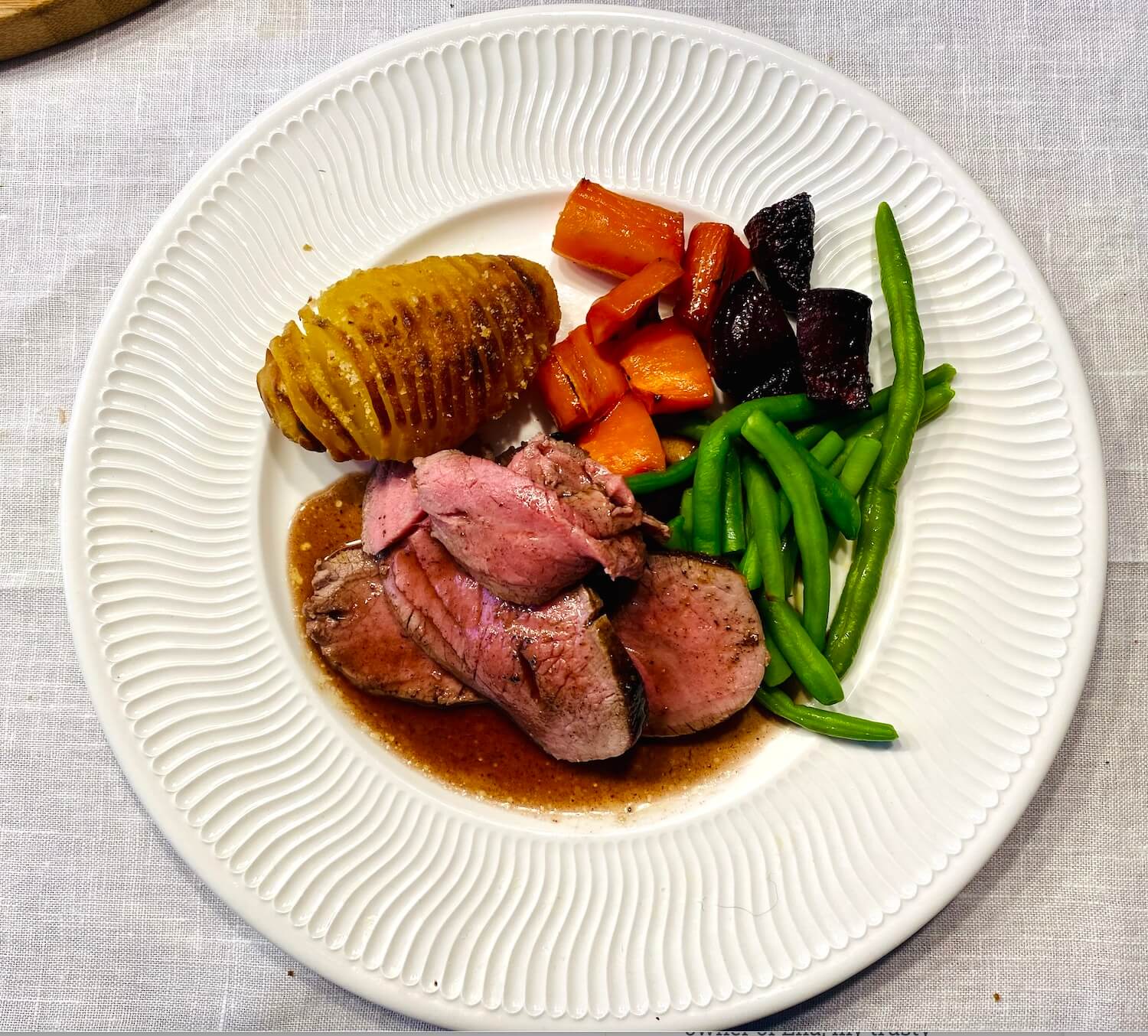 Sliced venison roast with red wine and juniper sauce, served with Hasselback potatoes, green beans, roasted beets, and carrots on a white plate.