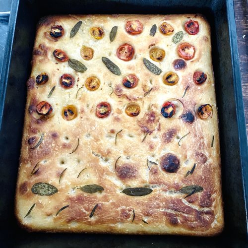Golden-brown sourdough focaccia topped with cherry tomatoes, rosemary, and sage on a baking dish.