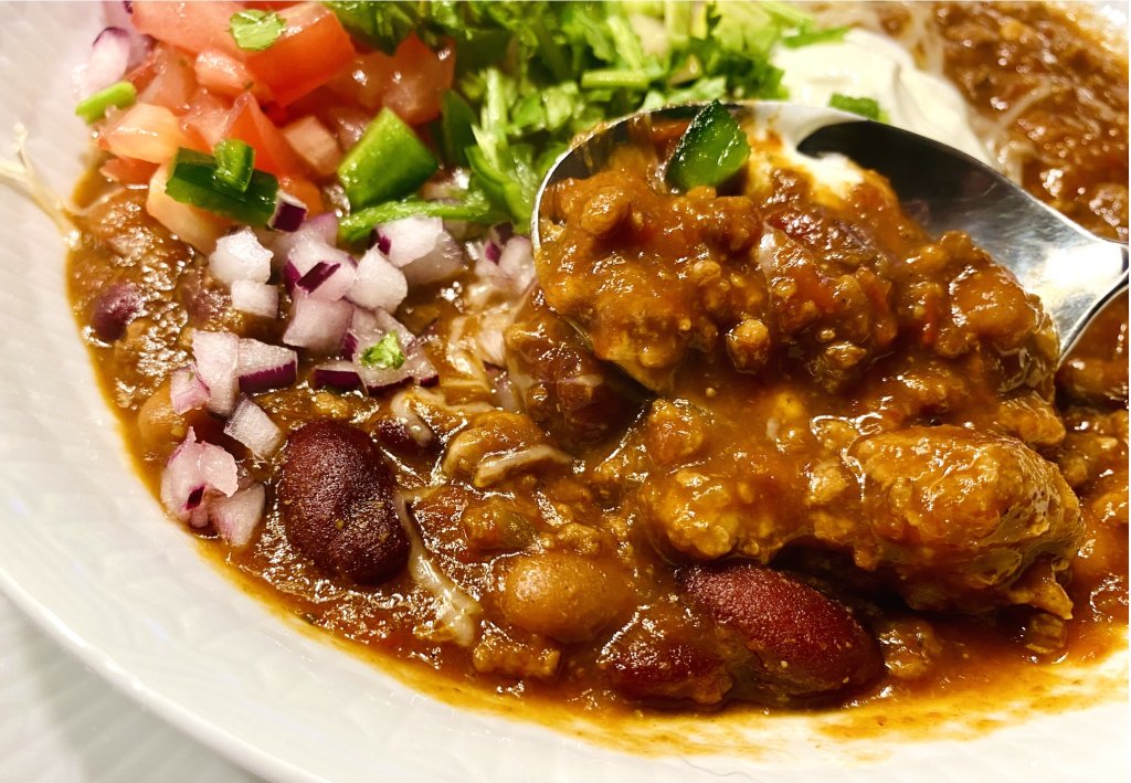 Close-up of a spoonful of spicy beef chili with beans, topped with fresh cilantro, tomato, avocado, sour cream, and cheese.