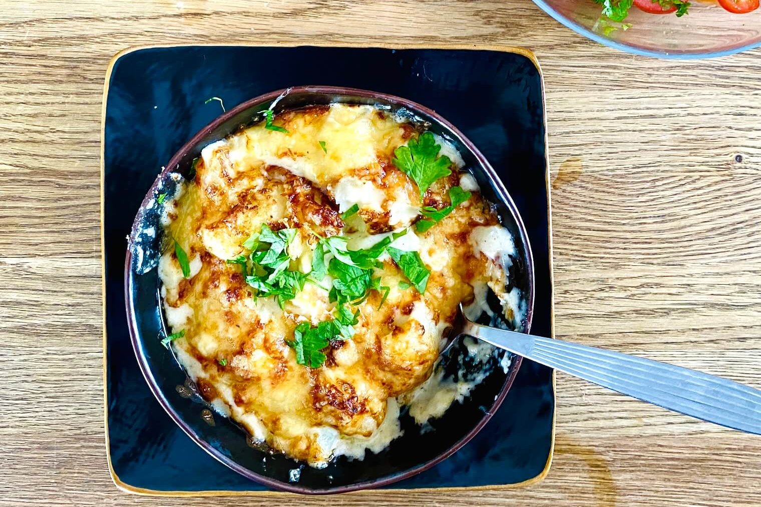 Finnish cauliflower gratin in a rustic bowl on a wooden table, garnished with parsley, with tomato salad partially visible in the background.