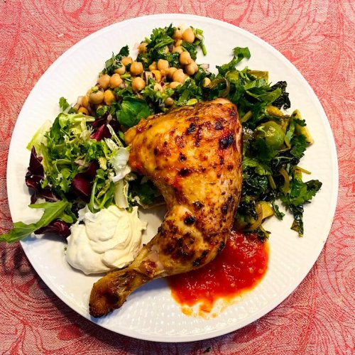 A golden roasted chicken thigh on a plate served with hot sauce, chickpea salad, fresh green salad, and roasted green vegetables.