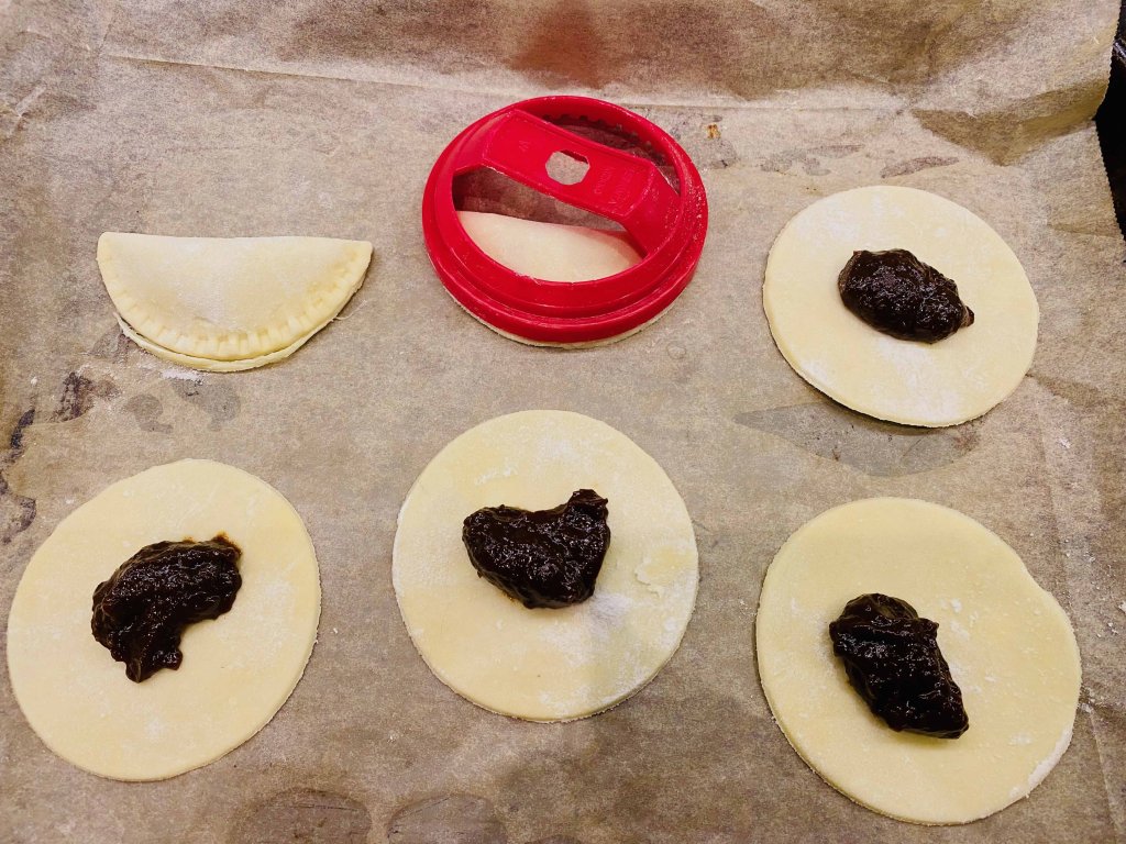 Several raw joulutortut on a tray, with plum filling in the centers. One tart is already filled and sealed, and the pastry cutter used to shape the rounds is visible beside them.