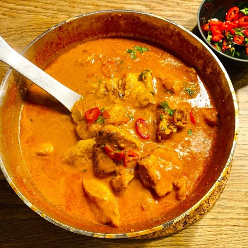 Butter chicken in a saucepan with roasted chicken pieces, fresh herbs, and red chili slices.