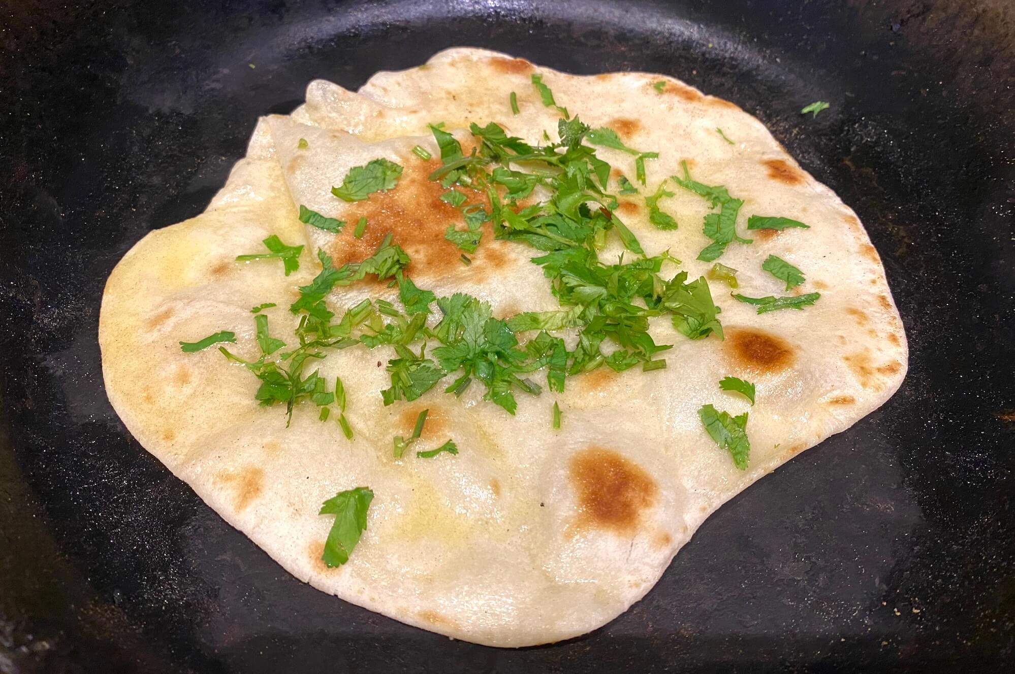 Flatbread cooking in a skillet, brushed with garlic butter and topped with fresh herbs.