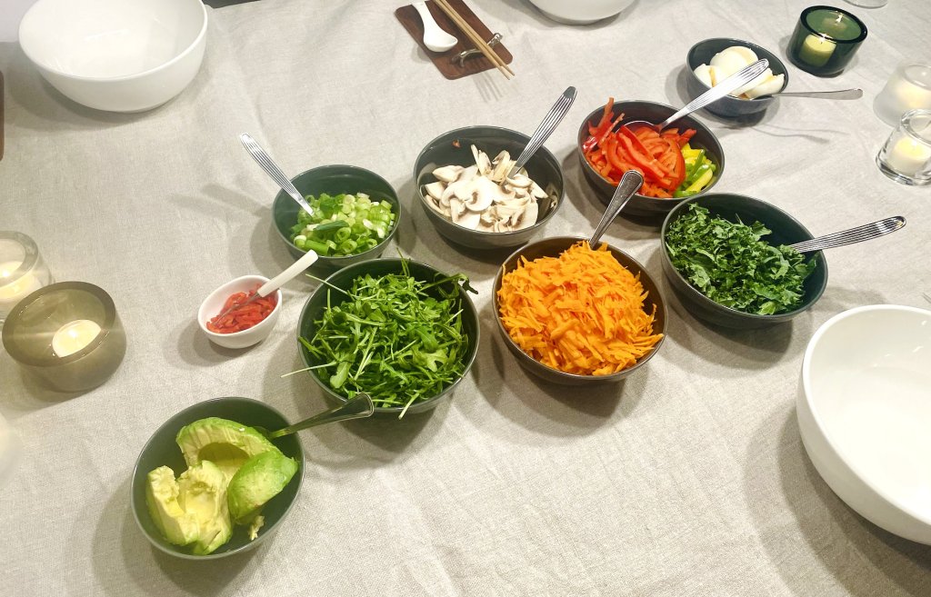 Variety of ramen toppings in individual bowls, including soft-boiled eggs, vegetables, avocado, and fresh herbs.