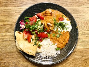 Plate of red lentil dahl with butter chicken, basmati rice, raita, folded flatbread, and tomato-cucumber salad