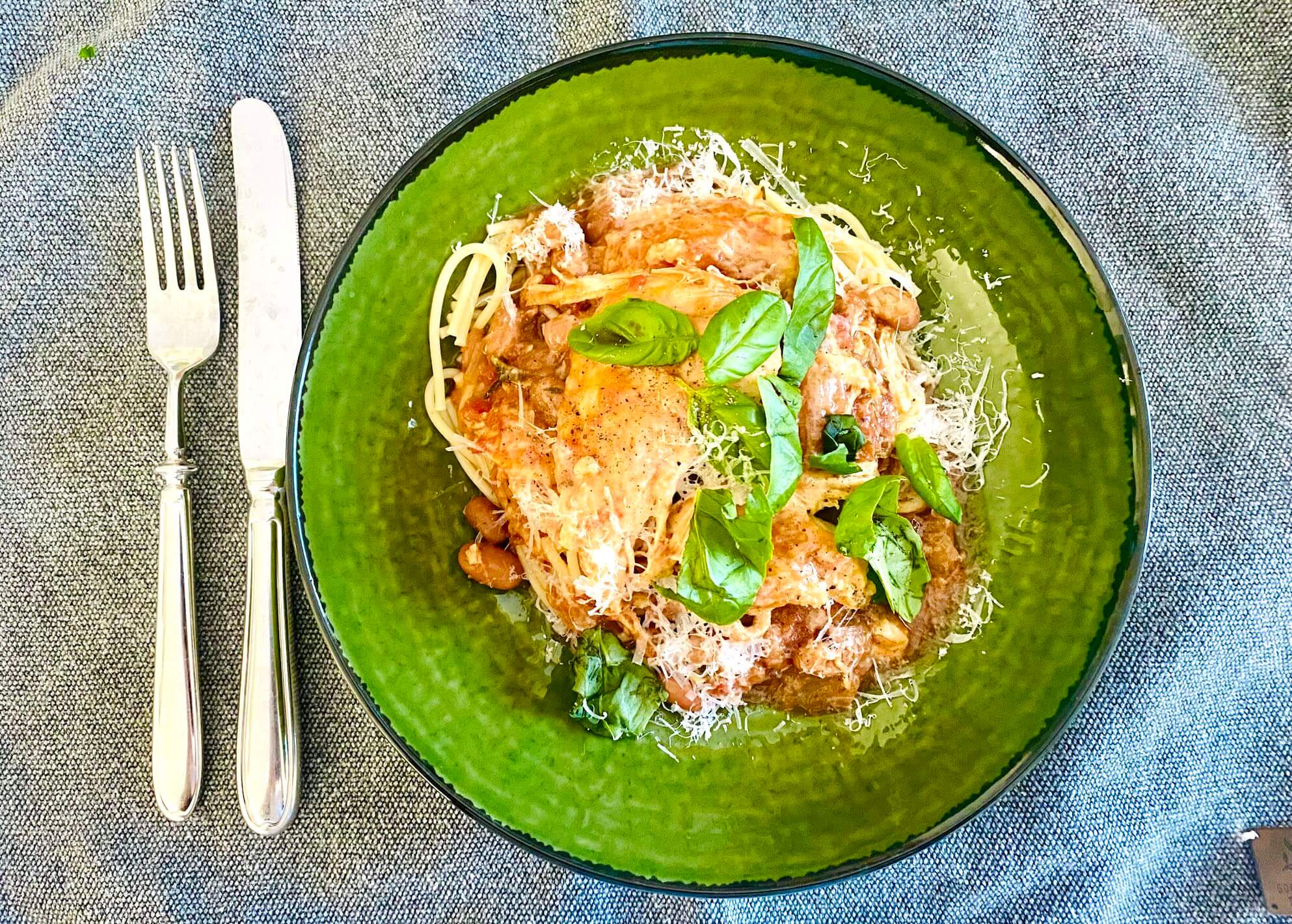 Chicken with Olives and Borlotti Beans served over spaghetti, topped with grated Parmesan and fresh basil, on a rustic green plate.