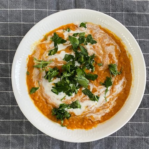 Bowl of spiced red lentil soup garnished with a swirl of Greek yogurt and chopped fresh parsley, served in a rustic bowl on a wooden surface.