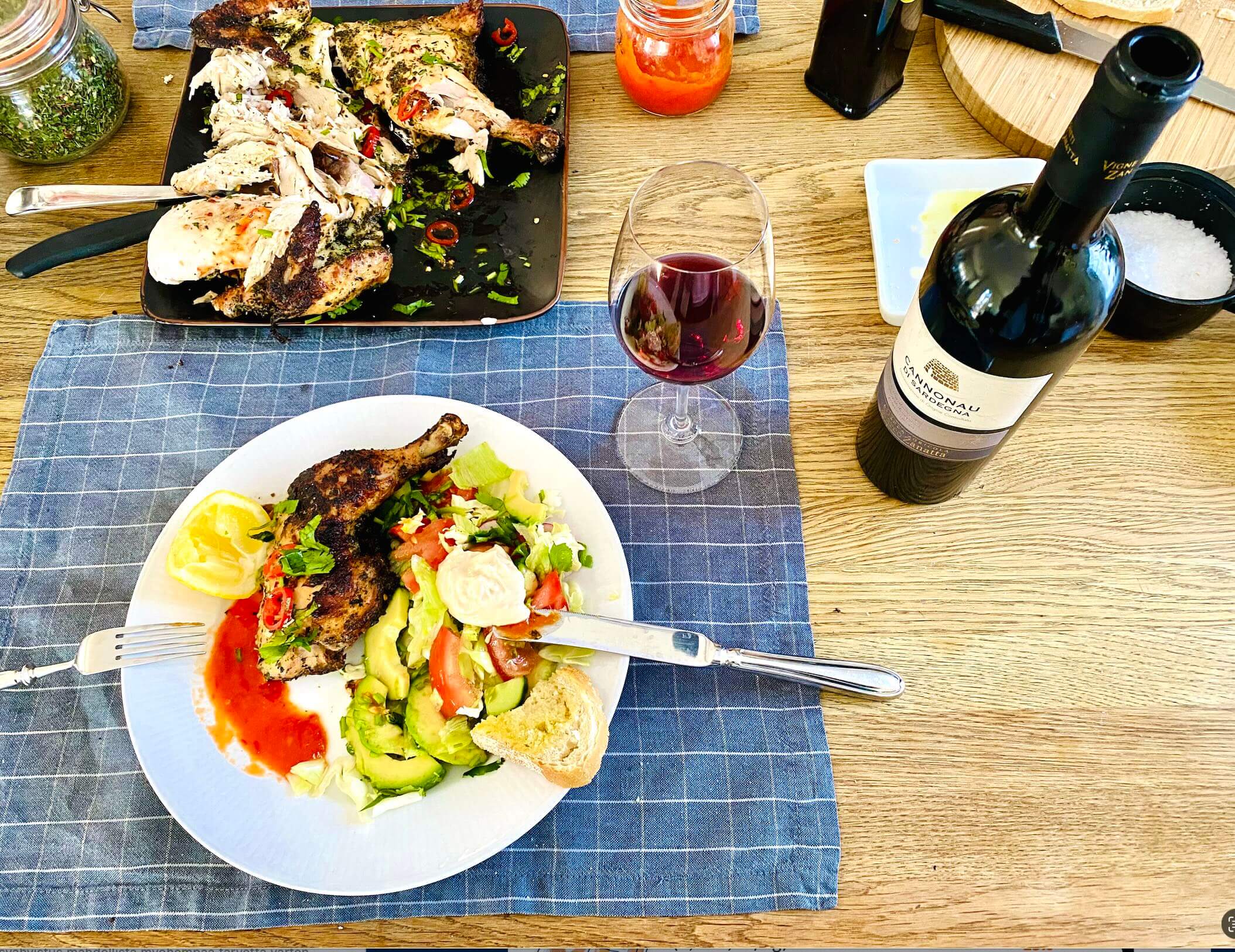 Grilled spatchcock chicken served with salad, rustic bread dipped in olive oil, homemade hot sauce, and red wine on a table setting